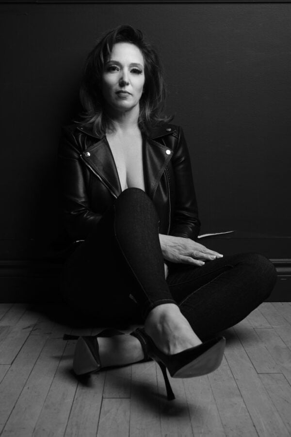 B&W photo of woman sitting down on the floor in a leather jacket and jeans with black heels